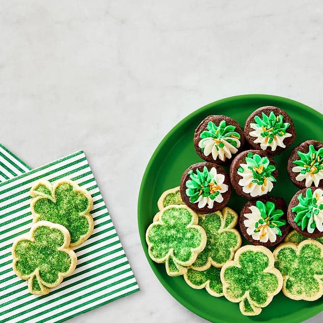 Green shamrock cookies are shown on a green stripe napkin & green plate. Along with mini brownies topped with green icing.
