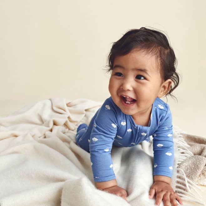 Smiling baby in a blue cloud print sleeper crawling on a soft cream blanket.