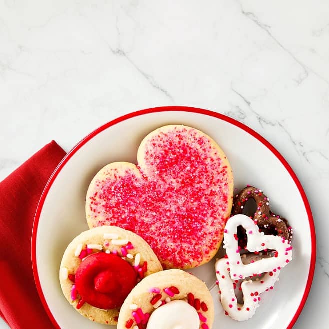 A plate of cookies is shown, heart-shaped, pink & sprinkles.