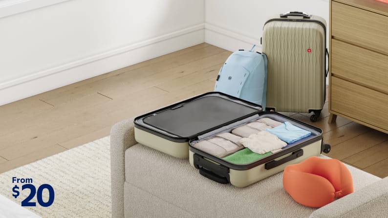 An opened hardshell suitcase displays neatly packed clothing and lays next to a neck pillow, backpack, and an additional matching suitcase.