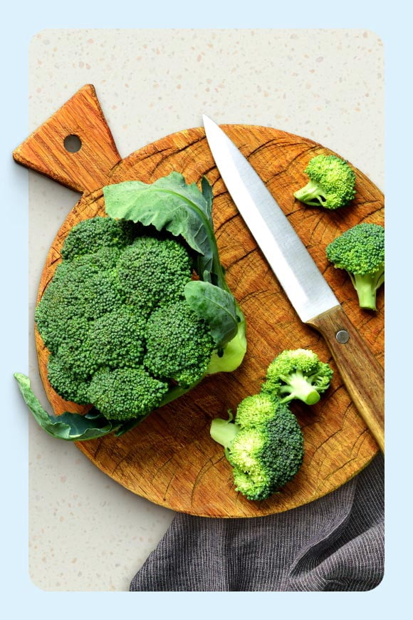 A head of broccoli has been cut up into pieces on a wooden cutting board with a knife. The board is on a marble counter.