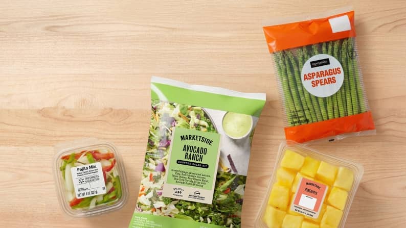An assortment of packaged produce sits on a kitchen table, including a salad kit, green beans, pineapple chunks and diced peppers.