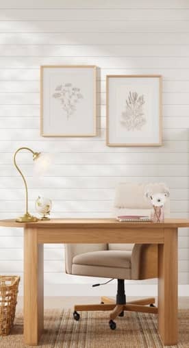 A light wood office desk is paired with a beige chair, a gold floral desk lamp and framed floral prints on the wall.
