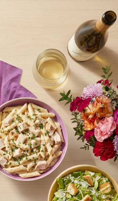 A tablescape of chicken penne alfredo, Ceasar salad, Josh white wine, and fresh pink and orange flowers is shown.