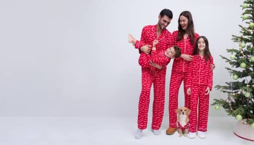 Family of four in matching red holiday pajamas with a small dog in front beside a decorated Christmas tree.