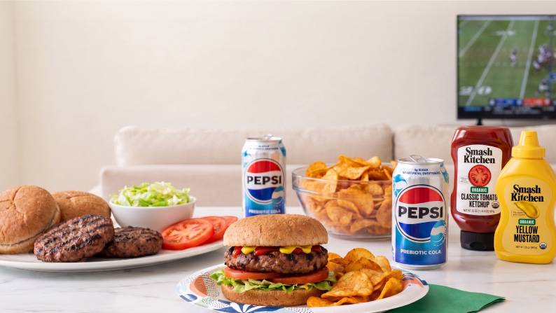 A paper plate with a hamburger and chips on a counter with a serving tray of burgers, buns and toppings, cans of Pepsi, chips, ketchup and mustard.