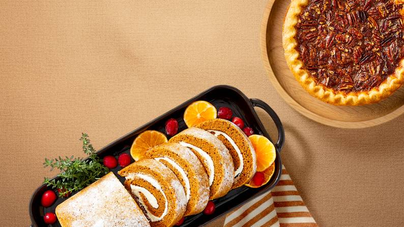A sliced pumpkin roll is shown on a platter garnished with cranberries and orange slices next to a whole pecan pie.