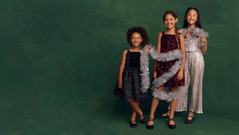 A girl wearing a black dress, a girl wearing a sequin red dress, and a girl wearing a silver dress hold an ornament in front of a green background.