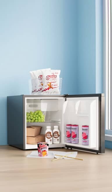 A small fridge is open on the floor with drinks, grapes and take out containers inside. Cheetos are on top of the fridge and Buldak is on the floor.