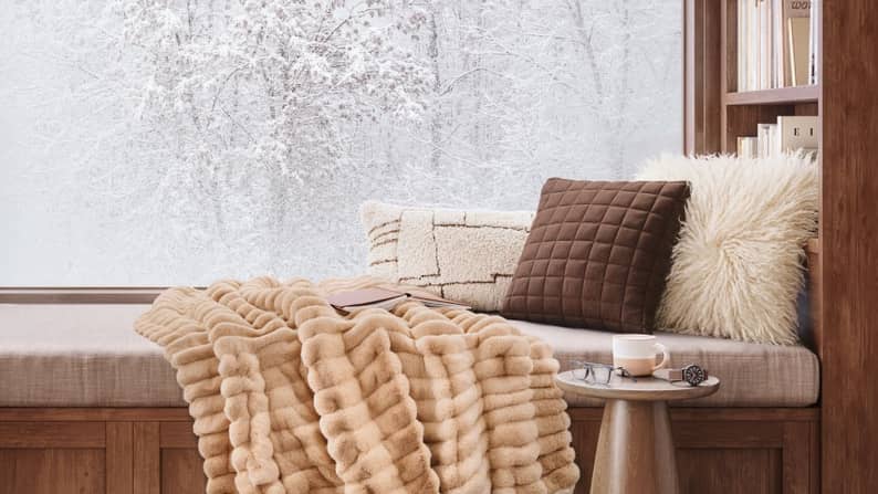 Cozy window seat with plush pillows, thick tan throw blanket, side table with cup and saucer, and snowy trees outside.