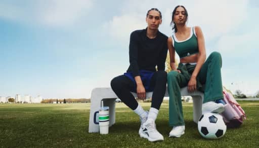 A young man and a young woman with her foot on a soccer ball wearing stylish activewear are sitting on a cement bench outside.