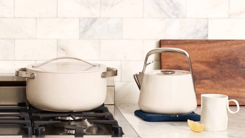 A lidded pot on a gas stovetop and kettle on a marbled countertop. A wood cutting board and a cup of tea with a lemon wedge rests on the counter.
