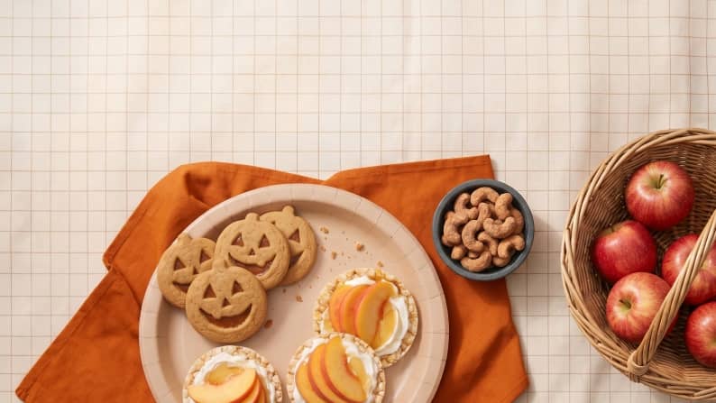 A plate with jack-o'-lantern cookies and rice cakes topped with sliced apples next to a bowl of seasoned cashews and a basket of apples.