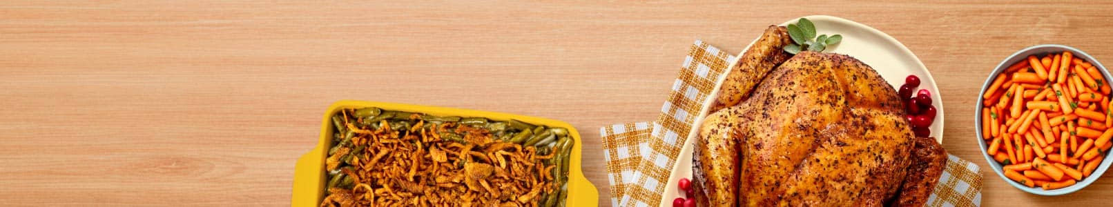 Image shows a green bean casserole in a baking dish, a turkey garnished with cranberries on a checked cloth and a bowl of carrots on a wooden table.