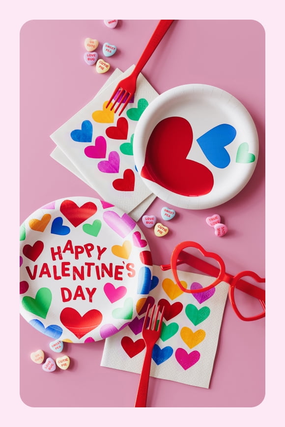 Multi-color Valentine’s Day paper plates, napkins, and red cutlery are scattered on a pink background.