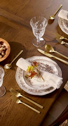 Gold-tone flatware, floral dinnerware, glassware, and a rolled-up napkin are set on a dark wooden dining table.