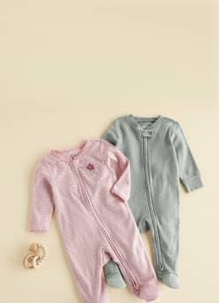 Two long sleeve zip front baby footed sleepers, one striped and one solid, with a wooden teether beside them.