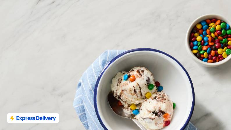 Two bowls of chocolate and vanilla ice cream topped with candy, next to waffle cones and candy toppings.