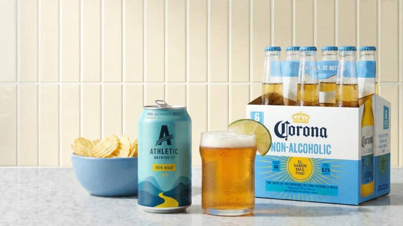 A bowl of rippled potato chips next to a can and glass of Athletic Brewing Company Run Wild IPA and a six pack of Corona non-alcoholic beer.