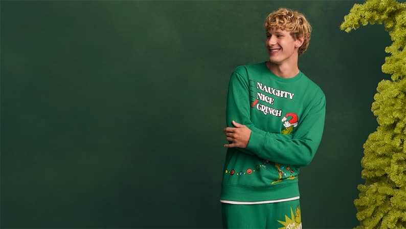 A man wearing a green Grinch set is pictured on a green backdrop next to a whimsical fake Christmas tree.