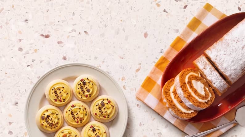 A pumpkin roll with two slices cut out next to a plate of sugar cookies with yellow icing and multicolored sprinkles.