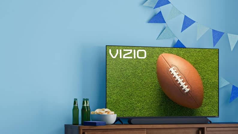 A big screen TV displays a football on the field with a flag banner behind it and snacks on the TV stand