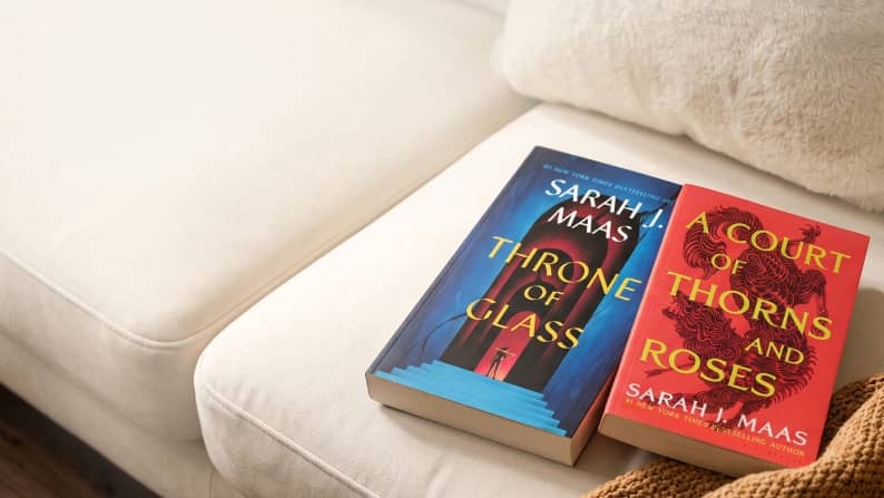 Sarah J. Maas fantasy novels Throne of Glass and A Court of Thorns and Roses on a cream sofa beside a textured blanket.