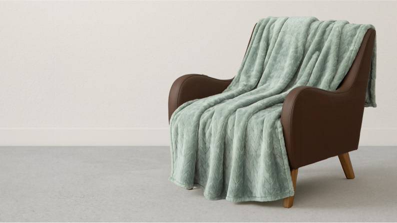 A sage green throw blanket is shown draped over a brown accent chair in a room with gray walls.