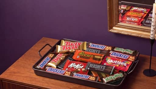 Tray of assorted Halloween candy bars and treats including Twix, Snickers, Reese’s, Hershey’s, Skittles, KitKat and Milky Way on a table.