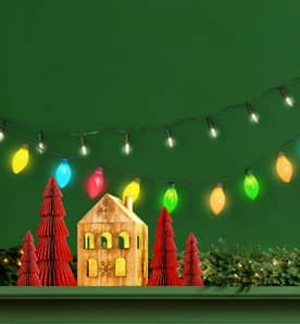 Image shows a close-up of a mantel decorated with Christmas house figurines, reindeer and large multi-colored Christmas lights.