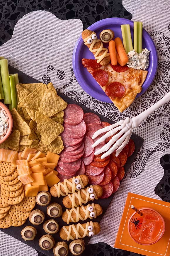 A counter full of sliced meat and cheese, cut vegetables, crackers, dip, an orange drink, and a plate with a slice of pizza and veggies with dip.