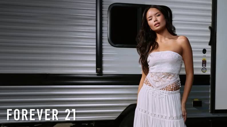 Young woman standing by an RV in a white sheer lace strapless tube top and white tiered maxi skirt. Forever 21 logo is seen.