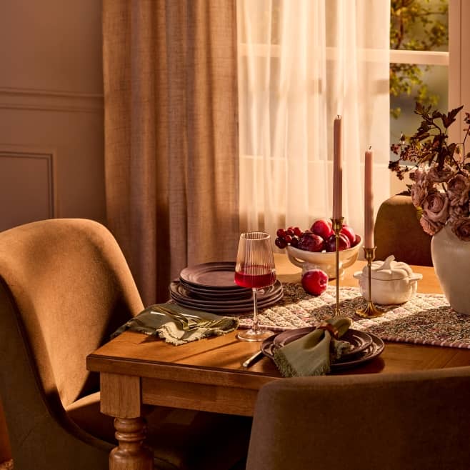 Warm dining room with a wooden table set with plates, glassware, candles and a vase of dried flowers.