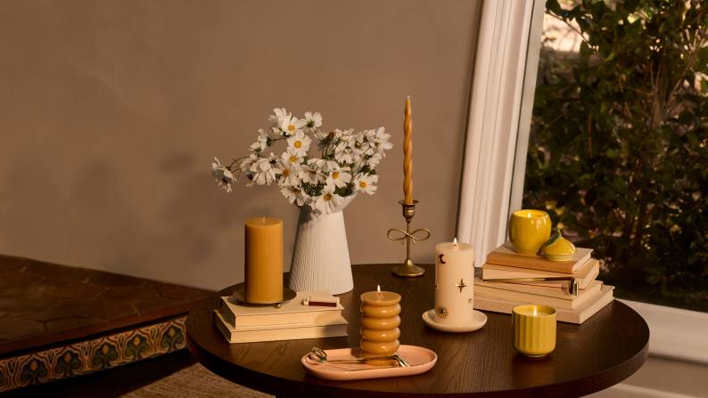 Yellow and white candles in various shapes and holders are styled on a dark wood table with white flowers and a stack of books.