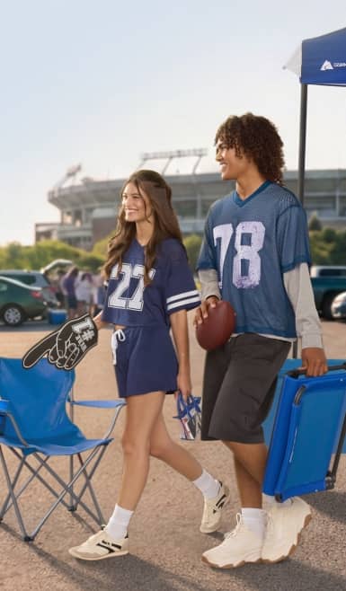 Two people in a stadium parking lot wearing jerseys. Two chairs.