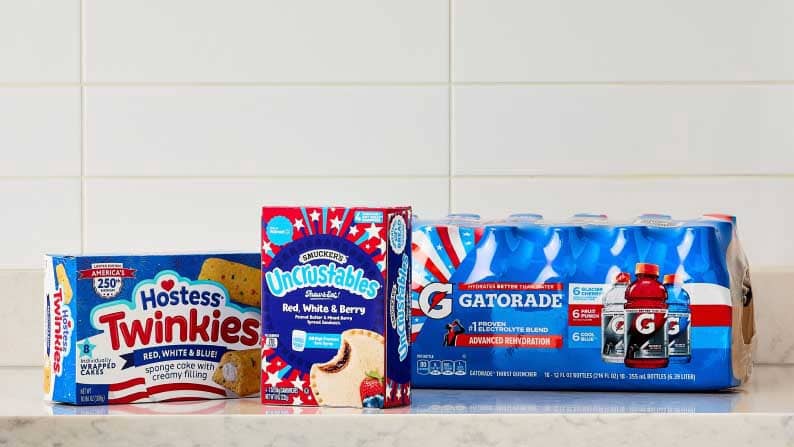 Hostess red, white, and blue Twinkies, Smucker's red, white, and berry Uncrustables, and a case of Gatorade on a marble counter.