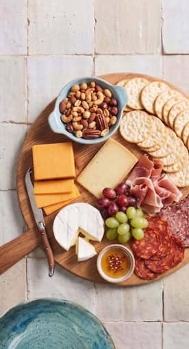 A wooden platter with brie, cheddar, salami, prosciutto, crackers, grapes, honey, and mixed nuts on a tile counter.