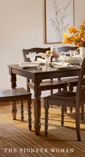 A dark wooden dining table with matching chairs is set with dinnerware, flatware, stemware and decor. Art hangs on a cream wall.