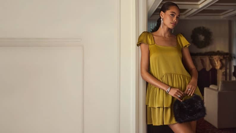 A woman wearing a yellow dress with bows at the shoulders is pictured standing in a home with a wreath on the wall.