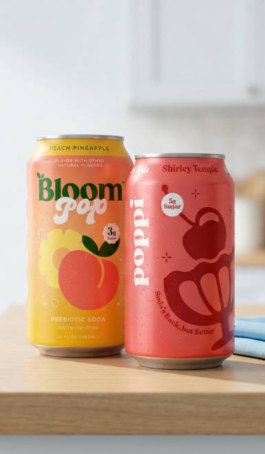 Two cans are shown on a wooden countertop: peach pineapple Bloom Pop and Shirley Temple Poppi.
