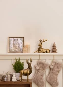 Gold reindeer figurines, owl art, small trees, wrapped gifts and two fluffy stockings decorate a cozy holiday mantel.
