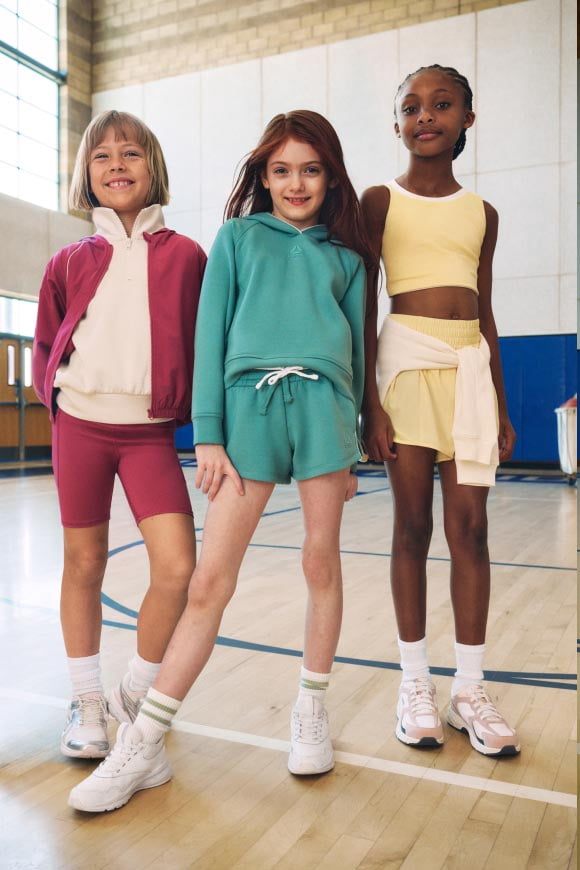 Three girls standing on a gym floor wearing colorful activewear outfits and sneakers.