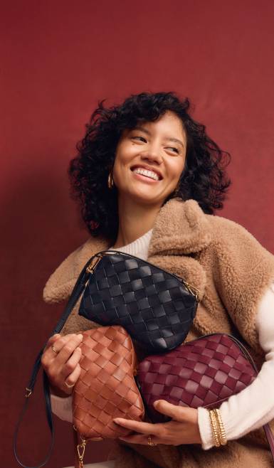 An image of a model smiling while holding black, brown and burgundy bags.