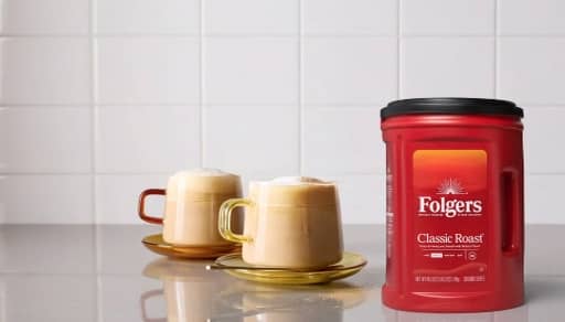 A canister of Folgers classic roast is shown on a counter next to two mug of coffee on saucers.