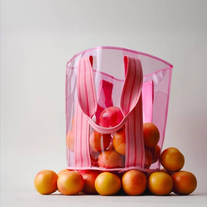 A clear beach tote with pink straps with oranges inside is shown.