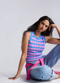 Woman sitting in a blue-and-pink striped sleeveless knit top and jeans, with a pink sweater tied at the waist.