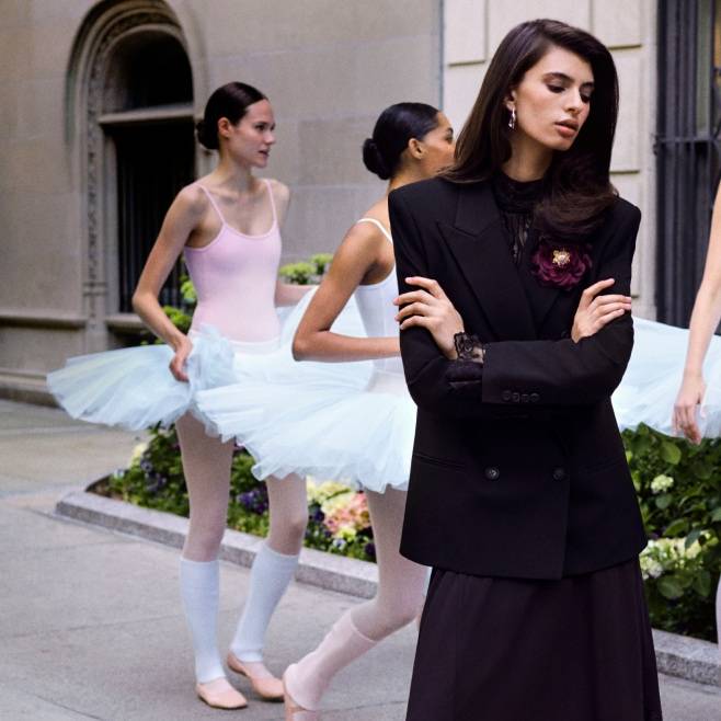 Woman on a sidewalk in a black tailored blazer over a black lace top and long black skirt with ballerinas in the background.