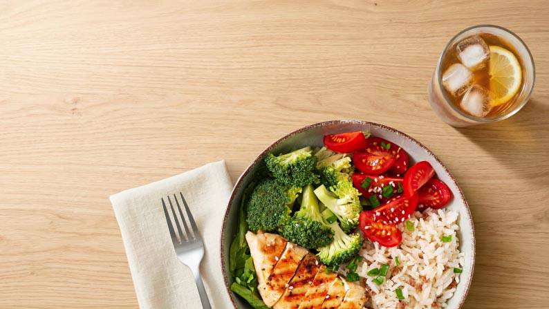 A bowl of broccoli, chicken, rice and tomatoes with a fork and napkin, next to a drink with lemon and ice in it.