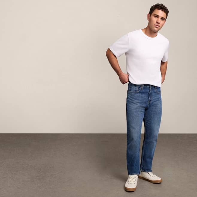 A man wearing straight leg jeans and white tee shirt is pictured on a beige backdrop.