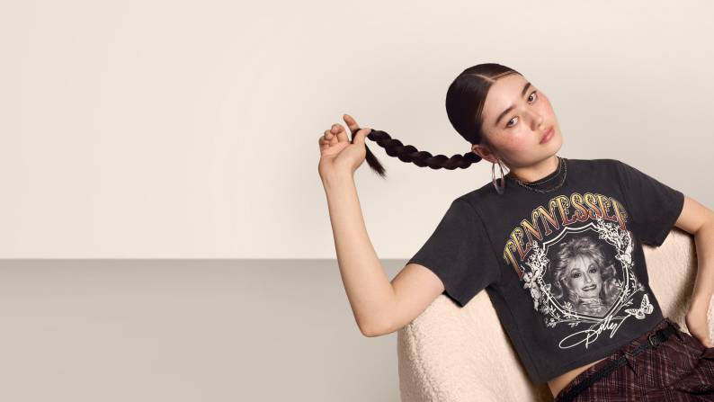A young woman wearing a Dolly Parton graphic tee shirt is pictured sitting a chair playing with her braid on a beige backdrop.